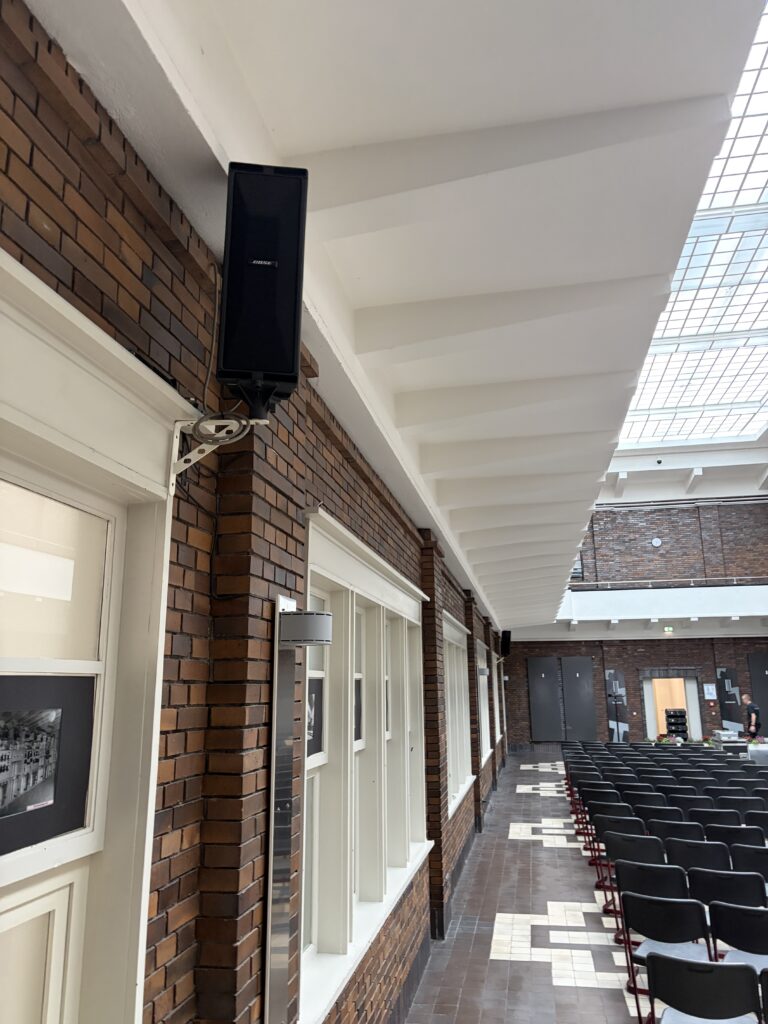 Wandmontierter Bose-Lautsprecher auf einer weißen Halterung an einer Backsteinwand in einem Veranstaltungssaal mit Reihenbestuhlung.